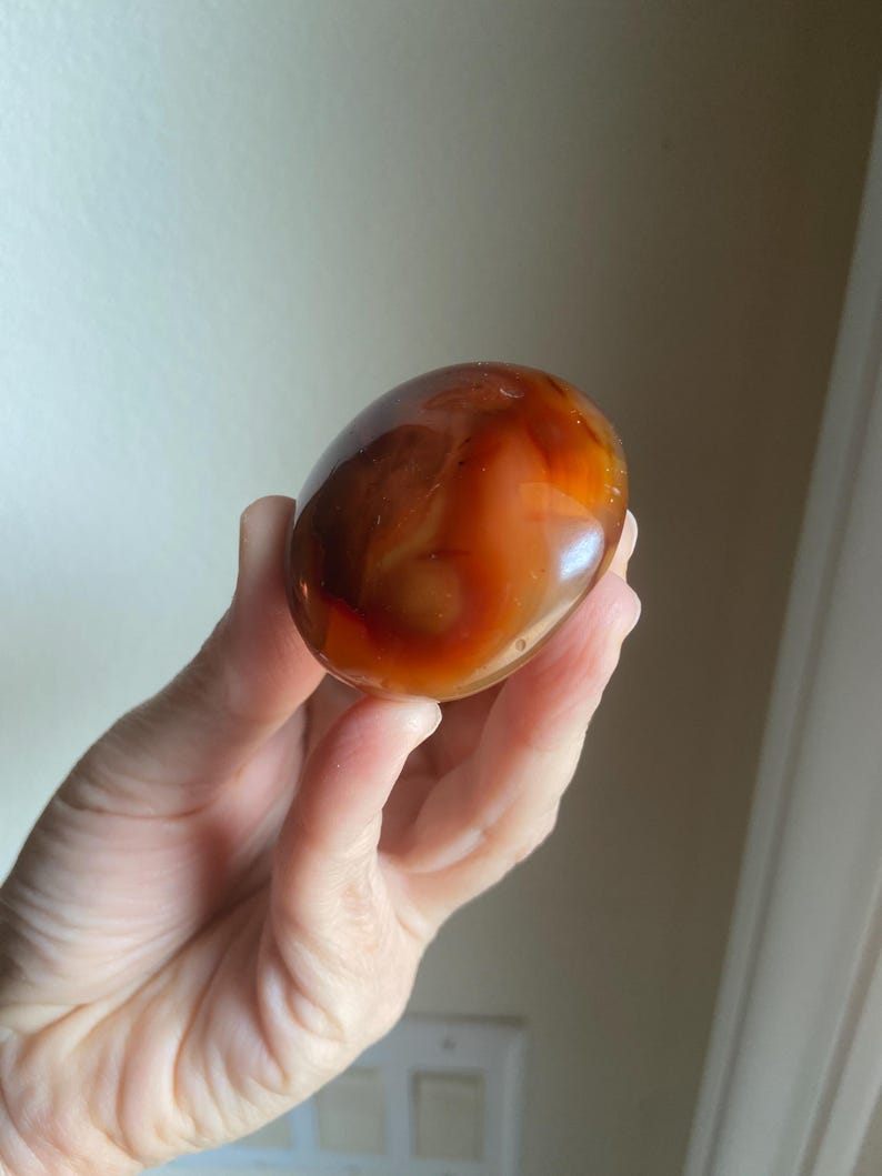 May include: A smooth, polished, oval-shaped carnelian stone featuring a translucent appearance with deep orange, reddish-brown, and amber color variations throughout its surface, displaying natural banding and a glossy, reflective finish.