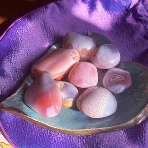 May include: A collection of polished gemstones in shades of pink, white, and orange, displayed on a leaf-shaped ceramic dish. The dish sits on a purple satin surface, with a drawstring bag visible in the background. The stones vary in size and pattern.