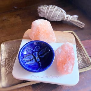 May include: A blue ceramic bowl with a dragonfly design, two pink Himalayan salt chunks, and a bundle of sage on a wooden and white tray. The tray has the text "One With Gems".