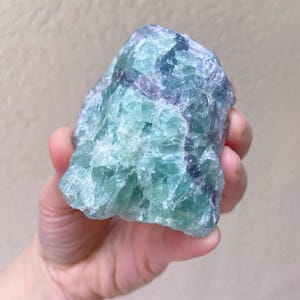 May include: A raw, natural fluorite crystal with a mix of blue, green, and purple hues. The stone has a rough, textured surface and is held in a hand against a neutral background. This mineral specimen is a unique decorative item.