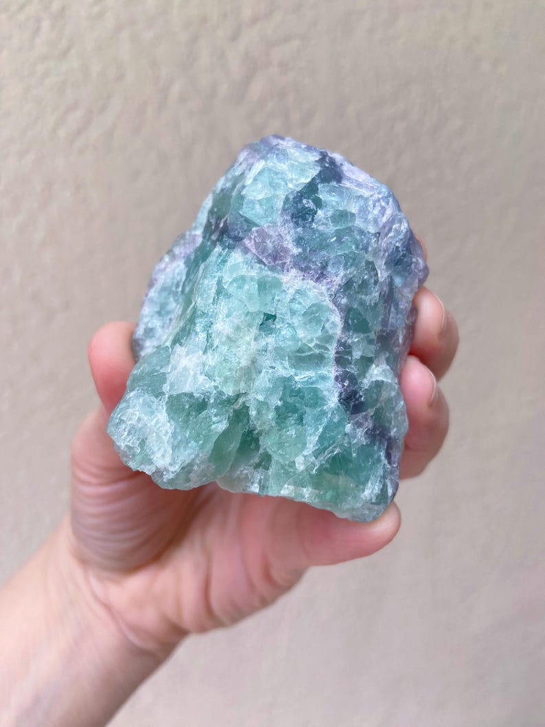 May include: A raw, natural fluorite crystal with a mix of blue, green, and purple hues. The stone has a rough, textured surface and is held in a hand against a neutral background. This mineral specimen is a unique decorative item.