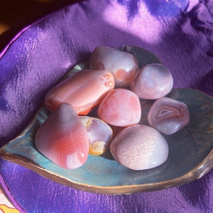May include: A collection of polished gemstones in shades of pink, white, and gray, displayed on a leaf-shaped ceramic dish. The stones vary in size and pattern, with visible banding and speckling. The dish sits on a purple satin surface.