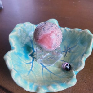 May include: A decorative ceramic leaf-shaped dish in shades of turquoise and green, holding a pink and gray sphere on a clear stand. A small, black and pink ladybug figurine rests on the dish's edge.