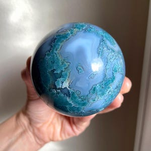 Pu&ograve; includere: Una sfera di pietra preziosa lucidata con un motivo a spirale di colori blu e verde acqua. La sfera &egrave; tenuta in una mano, mostrando la sua forma liscia e arrotondata. La superficie della pietra ha un aspetto marmorizzato, con aree di tonalit&agrave; pi&ugrave; chiare e pi&ugrave; scure.