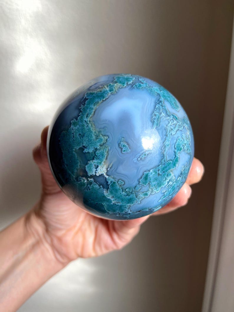 Pu&ograve; includere: Una sfera di pietra preziosa lucidata con un motivo a spirale di colori blu e verde acqua. La sfera &egrave; tenuta in una mano, mostrando la sua forma liscia e arrotondata. La superficie della pietra ha un aspetto marmorizzato, con aree di tonalit&agrave; pi&ugrave; chiare e pi&ugrave; scure.