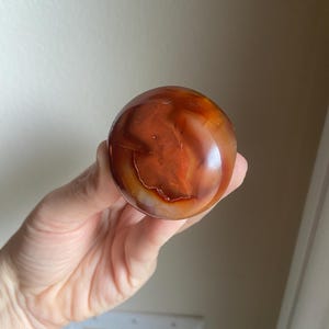 May include: A polished, spherical carnelian gemstone displaying a vibrant range of deep orange, reddish-brown, and translucent white banding. The stone features a smooth, glossy surface with natural internal inclusions and varied color gradients.