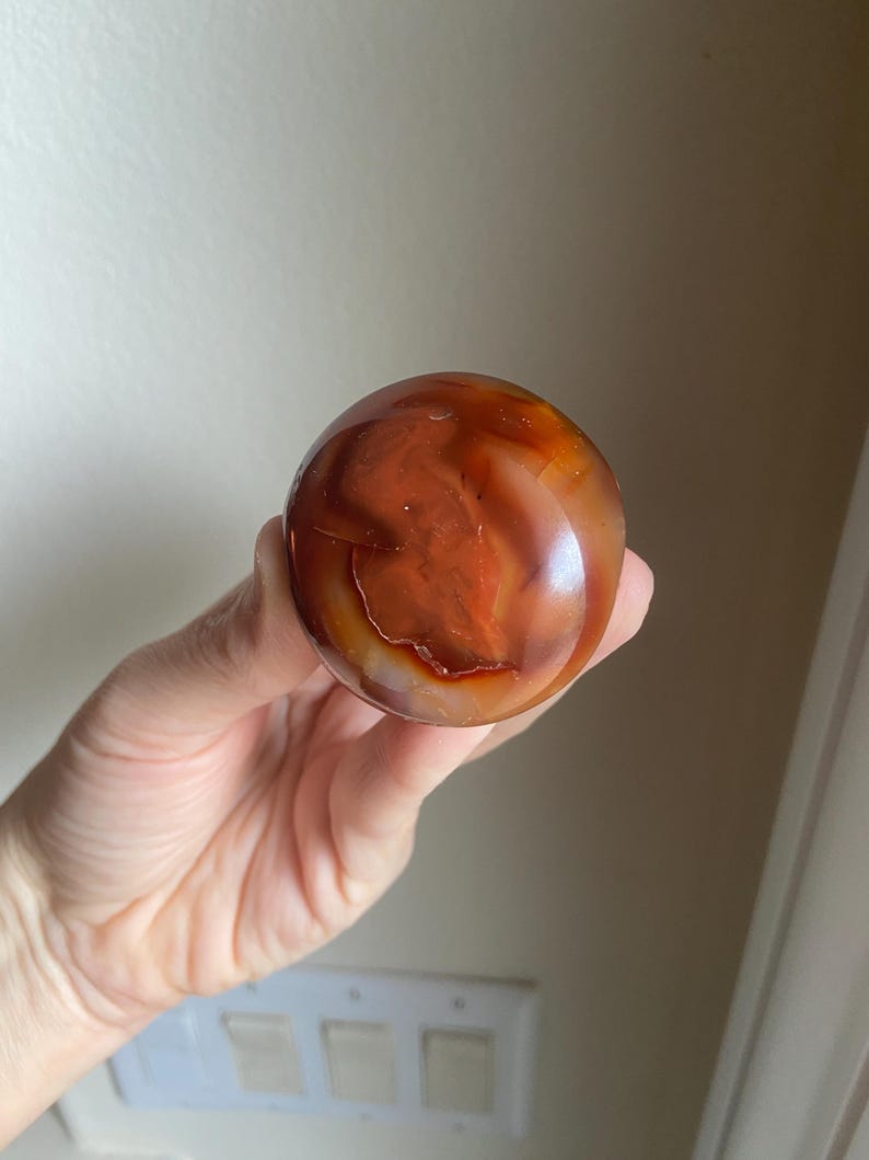 May include: A polished, spherical carnelian gemstone displaying a vibrant range of deep orange, reddish-brown, and translucent white banding. The stone features a smooth, glossy surface with natural internal inclusions and varied color gradients.