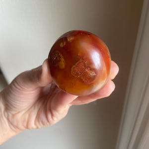 May include: A polished, spherical carnelian stone displaying a warm palette of deep orange, burnt red, and translucent amber tones. The surface features natural inclusions and organic, cloud-like patterns with a smooth, glossy finish throughout.