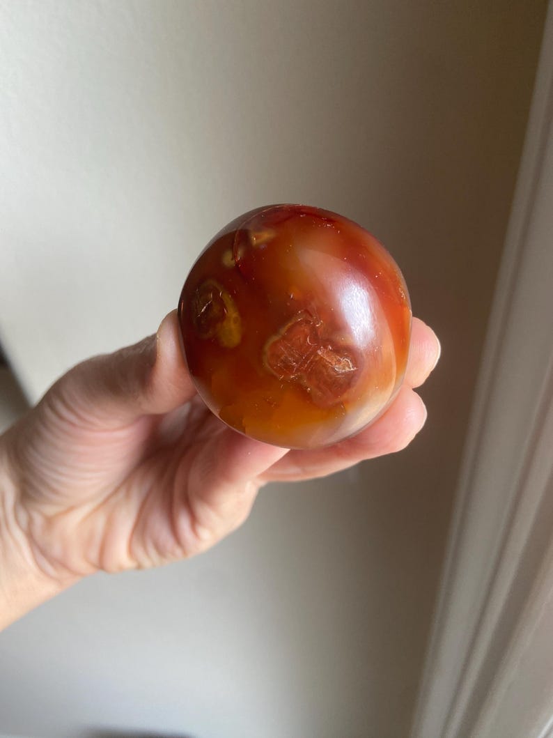 May include: A polished, spherical carnelian stone displaying a warm palette of deep orange, burnt red, and translucent amber tones. The surface features natural inclusions and organic, cloud-like patterns with a smooth, glossy finish throughout.