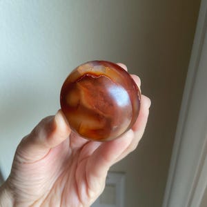 May include: A polished, spherical carnelian gemstone displaying a rich gradient of deep burnt orange, reddish-brown, and translucent amber tones. The surface features natural swirling patterns and internal inclusions that create a smooth, glossy appearance.