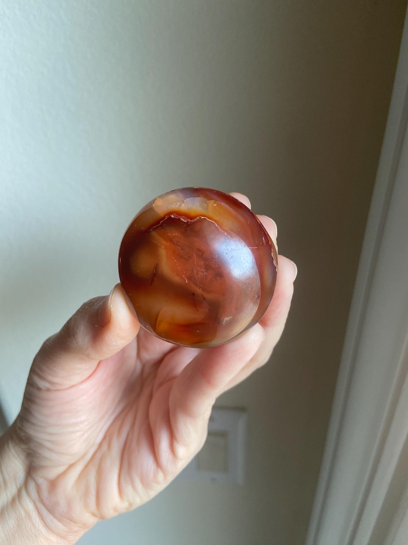 May include: A polished, spherical carnelian gemstone displaying a rich gradient of deep burnt orange, reddish-brown, and translucent amber tones. The surface features natural swirling patterns and internal inclusions that create a smooth, glossy appearance.