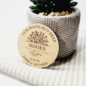 May include: A circular wooden keepsake for a family reunion, featuring the text "OUR ROOTS RUN DEEP HOOKS FAMILY REUNION 9.14.24". The wooden disc has a tree design and is placed in front of a small potted succulent plant.