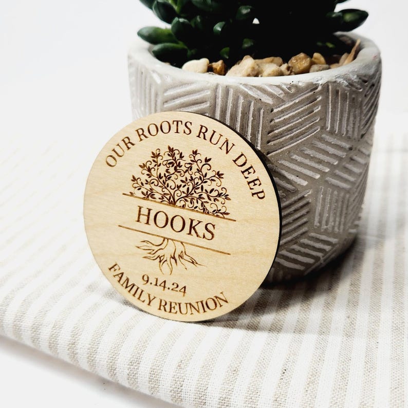 May include: A circular wooden keepsake for a family reunion, featuring the text "OUR ROOTS RUN DEEP HOOKS FAMILY REUNION 9.14.24". The wooden disc has a tree design and is placed in front of a small potted succulent plant.