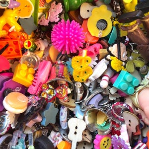 May include: A vibrant assortment of small, colorful trinkets and charms. The collection includes various shapes like stars, flowers, and musical instruments, along with toy blocks, buttons, and other decorative items. The items are in a variety of colors, including pink, yellow, green, and blue.