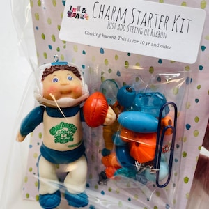 May include: A clear plastic bag contains a Cabbage Patch Kids charm starter kit. The kit includes a Cabbage Patch Kids doll ornament in a football uniform holding a football, and other colorful beads. The text on the label reads "CHARM STARTER KIT".