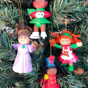 May include: Four Cabbage Patch Kids Christmas ornaments hanging on a tree. One is an ice skater in red and green, one is an angel in a lavender gown with a trumpet, one has red yarn hair in a red dress, and one is a soldier in a red uniform.