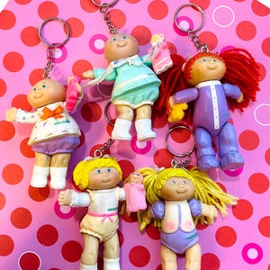 May include: Five Cabbage Patch Kids keychains featuring small plastic dolls with yarn hair and colorful outfits. Each doll is attached to a silver metal keyring and chain. The dolls have various hair colors including red and yellow, and wear outfits.