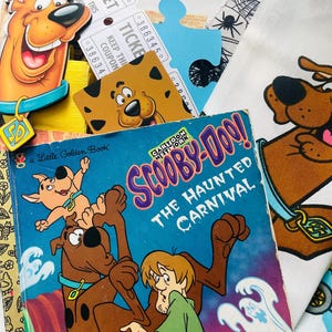 May include: A collection of Scooby-Doo themed items including a Little Golden Book titled Scooby-Doo! The Haunted Carnival, a die-cut Scooby-Doo character cutout, two paper tickets, a brown patterned card, a blue puzzle piece, and white fabric.