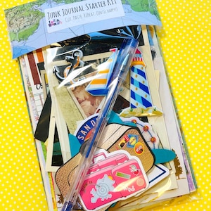 May include: A clear plastic bag filled with junk journal supplies. The kit includes decorative stickers of suitcases, a lighthouse, and an anchor. A blue pen rests on top of the ephemera. The bag is labeled "JUNK JOURNAL STARTER KIT".