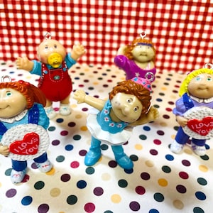 May include: Five Cabbage Patch Kids figurines. Each doll is in a different pose and outfit, with details like a pacifier, overalls, and heart-shaped signs that read "I Love You". The background is a red and white checkered pattern.