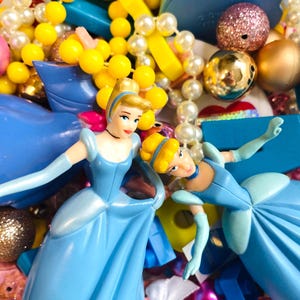 May include: A vibrant assortment of toys and beads. Two Cinderella figurines in light blue dresses are the focal point, surrounded by yellow and white beads, and other decorative elements. The scene is filled with a variety of textures and colors.