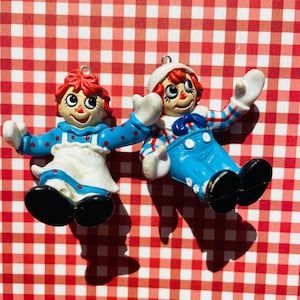 May include: Two Raggedy Ann and Andy doll ornaments. The dolls have red yarn hair, painted faces, and wear blue and white outfits. The ornaments are on a red and white checkered background.