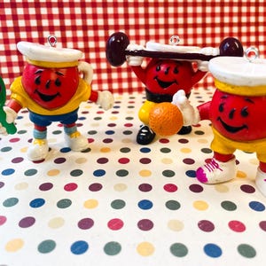 May include: Three vintage-style red, yellow, and blue figurines of a smiling character in various athletic poses. One holds a green paddle and purple ball, another lifts a barbell, and the third holds an orange ball. Each has a white hat.