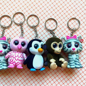 May include: A collection of five keychains featuring animal figurines. The keychains include a cat, owl, penguin, monkey, and another cat, each with a silver chain and ring. The cat keychains have pink bows.