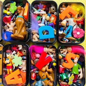 May include: A collection of small, black metal tins filled with various colorful toys and trinkets. The tins contain cartoon character figurines, plastic letters, flowers, and other decorative items. The background is a yellow surface with white polka dots.