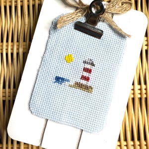Puede incluir: Un portapapeles de madera blanco con un lazo de cuerda marrón y un clip plateado que sujeta un punto de cruz azul y blanco de un faro con una torre de rayas rojas y blancas, un sol amarillo y una ola azul.