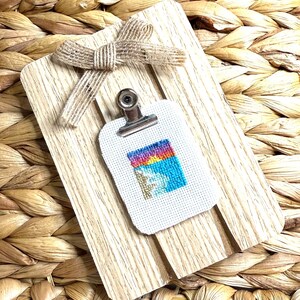 Puede incluir: Un pequeño lienzo blanco con un diseño de punto de cruz colorido de una puesta de sol sobre una escena de playa. El lienzo está sujeto a un portapapeles de madera con un lazo de arpillera marrón.