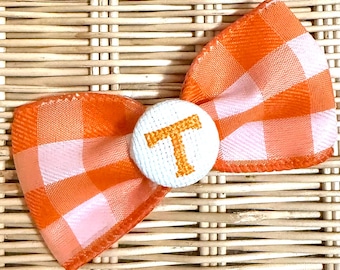 Könnte beinhalten: Orange-weißes Karoband mit einer weißen Knopfleiste mit einem goldenen gestickten Buchstaben "T".