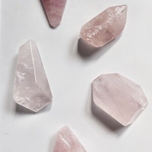 Pode incluir: Cinco cristais de quartzo rosa em vários formatos e tamanhos. Os cristais são lisos e polidos, com uma tonalidade rosa claro.