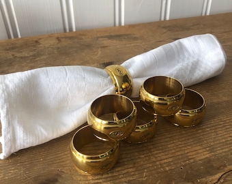 May include: Six gold-coloured napkin rings, each with a logo, displayed with a white linen napkin on a wooden table. The rings are circular and polished, reflecting light.
