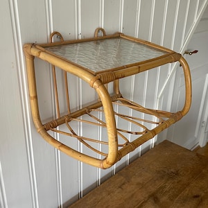 May include: A vintage wall-mounted shelf made of natural bamboo. It features a clear, textured glass top and a lower shelf with a woven design. The shelf is attached to the wall with two curved hooks. The bamboo has a warm, light brown color.