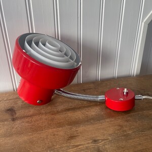 May include: A red vintage desk lamp with a flexible arm and a white, multi-layered shade. The lamp has a round base with a switch on the side.