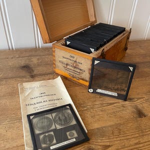 Könnte beinhalten: Eine Holzkiste mit Scharnierdeckel, gefüllt mit schwarzen Dias. Auf der Kiste steht schwedischer Text: "30 Skioptikonbilder Stockholm Historia". Neben der Kiste liegen zwei Dias und ein Heft mit demselben Text. Die Dias zeigen Bilder von Stockholm.