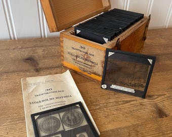 Op de afbeelding: Een vintage houten kist met een scharnierend deksel, met zwarte dia's. De kist is gemarkeerd met Zweedse tekst, "30 Skioptikonbilder Stockholm Historia". Naast de kist liggen twee dia's en een boekje met dezelfde tekst. De dia's tonen afbeeldingen van Stockholm.