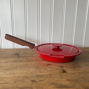 以下が含まれることがあります： 木製の取っ手とそれに合わせた蓋が付いた赤い鋳鉄製のフライパン。フライパンは木製の表面の上に置かれています。