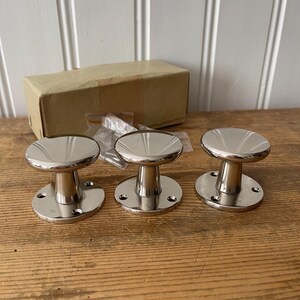 Op de afbeelding: Drie glanzende, zilverkleurige kastknoppen met een cirkelvormige basis en een afgeronde bovenkant. De knoppen staan op een houten oppervlak, met een kartonnen doos en een klein zakje schroeven op de achtergrond.