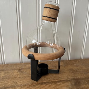 May include: A clear glass carafe with a wooden stopper and a wooden ring base. The carafe sits in a black metal stand with a small candle holder. The wooden elements have a natural finish.