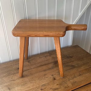 May include: A small wooden stool with a rectangular top and a handle extending from one side. The stool features three tapered legs and a natural light brown wood grain finish, showcasing a smooth surface and rounded edges on the top piece.