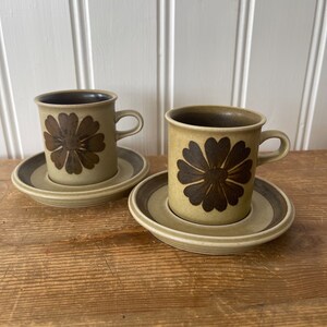 May include: Two brown and green ceramic coffee cups with a brown flower design. The cups are on matching saucers.
