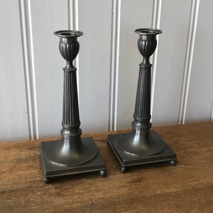 May include: Two antique pewter candlesticks with a square base and fluted column design. The candlesticks have a classic, elegant appearance, perfect for adding a touch of vintage charm to any room. They are displayed on a wooden surface.