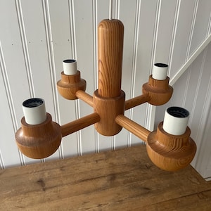 Può includere: Un lampadario in legno con cinque bracci che si estendono da una colonna centrale. Ogni braccio ha un portalampada con interno bianco. Il lampadario è realizzato in legno chiaro e ha un design moderno della metà del secolo.