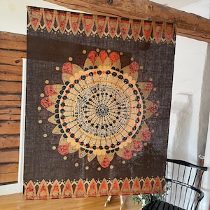 Puede incluir: Gran textil marrón oscuro con un diseño circular, similar a una flor, en dorado, naranja y negro. El diseño está centrado en la tela y enmarcado por un borde decorativo. El textil está colgado en una habitación.