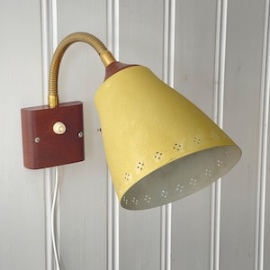 May include: A vintage wall-mounted lamp with a yellow conical shade and a flexible brass neck. The shade has decorative perforations. The lamp is attached to a wooden base with a switch and a white electrical cord.