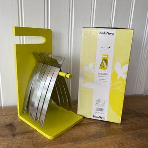 Könnte beinhalten: Ein gelber Kombi-Besteckhalter mit Edelstahlutensilien. Der Halter hat ein einzigartiges Design mit einem Griff und einem Schlitz für die Utensilien. Eine gelb-weiße Schachtel mit dem BodaNova-Logo befindet sich neben dem Halter.