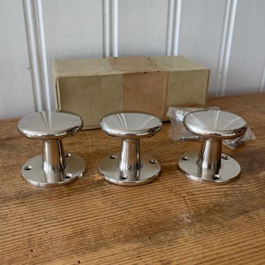 Può includere: Tre pomelli per mobili lucidi, color argento, con parte superiore e base circolari e stelo cilindrico. I pomelli sono esposti su una superficie di legno, con una scatola di cartone sullo sfondo. I pomelli sono nuovi.