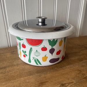 Puede incluir: Olla de esmalte blanco con tapa plateada y estampado de verduras de colores. El estampado presenta tomates rojos, judías verdes, pimientos amarillos, berenjenas moradas, una sandía y otras verduras.